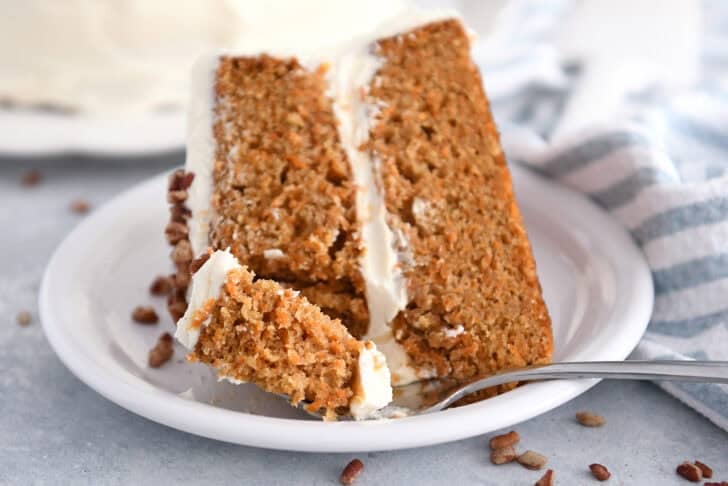 Bite of carrot cake on fork next to slice of frosted carrot cake on white plate.