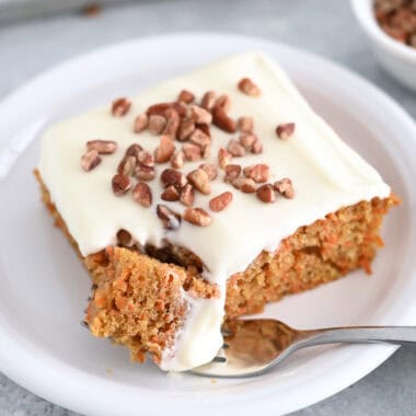 A piece of cream cheese frosted carrot sheet cake sprinkled with pecans on a white plate. A fork is taking a bite out of the cake.