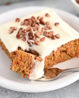 A slice of cream cheese frosted carrot sheet cake sprinkled with pecans on a white plate. Fork is taking a bite of cake.