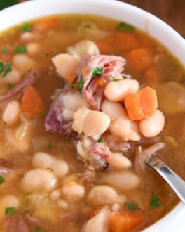 White bowl with handles with spoon lifting bite of white beans, carrots and ham.