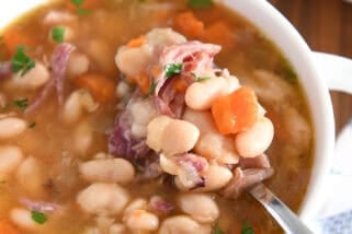 Top down view of ham and bean soup in white bowl with handles with spoon lifting bite of white beans, carrots and ham.