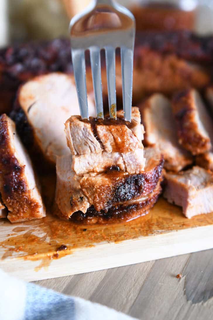 The fork pierced two stacked cooked pork tenderloin on a wooden cutting board.