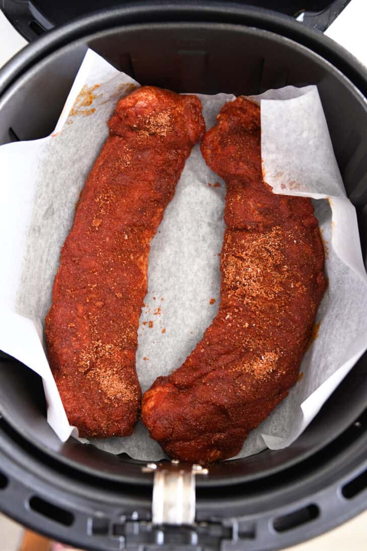 On the parchment paper in the air fryer, there are two uncooked pork tenderloin with spice shells.
