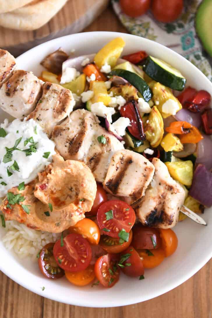 Upward down view of white bowl with chicken cubes, roasted pumpkin, zucchini, red onion and pepper, cherry tomatoes, rice, hummus and tzatziki sauce.