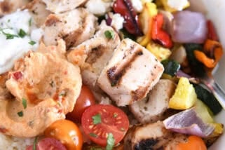White bowl with metal skewer with chicken pieces, roasted squash, zucchini, red onions and bell peppers, cherry tomatoes, rice, hummus and tzatziki sauce.