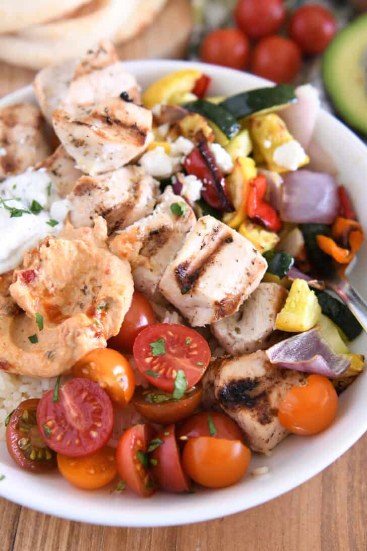White bowl with metal skewers, chicken cubes, grilled pumpkin, zucchini, red onion and pepper, cherry tomatoes, rice, hummus and tzatziki sauce.