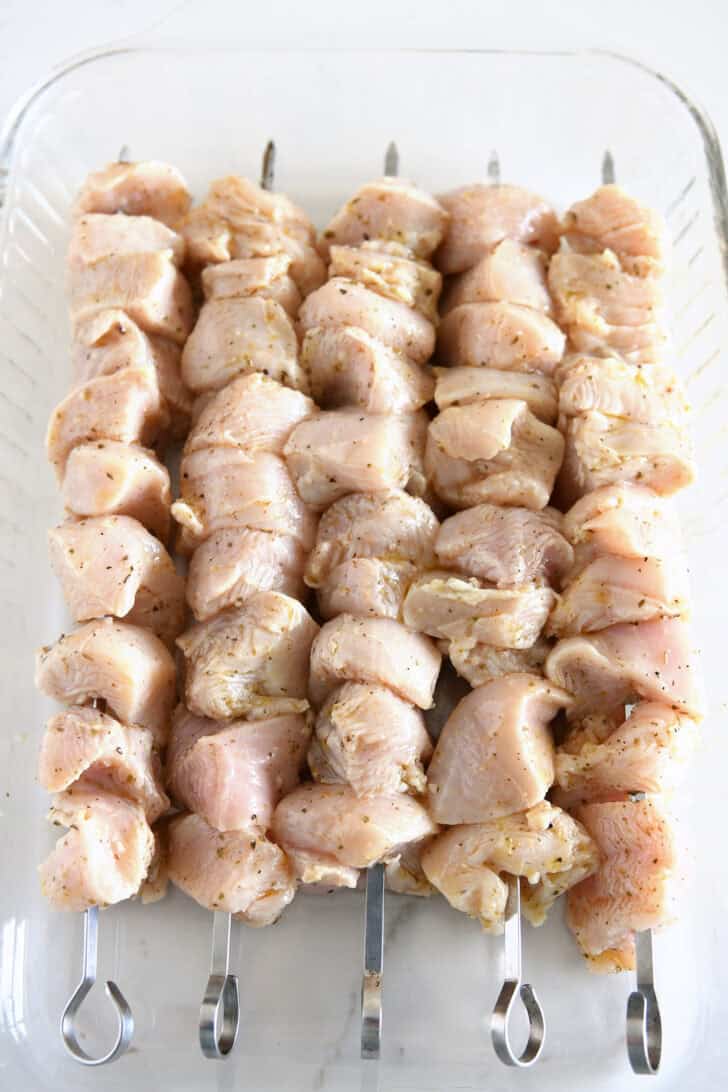 Glass 9x13-inch dish with raw chicken skewers with lemon marinade.