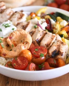 White bowl with metal skewer with chicken pieces, roasted squash, zucchini, red onions and bell peppers, cherry tomatoes, rice, hummus and tzatziki sauce.