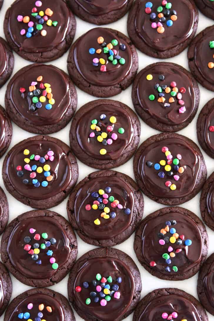 Top-down view of many chocolate chip cookies, topped with Ganache and sprinkled with rainbow chips.
