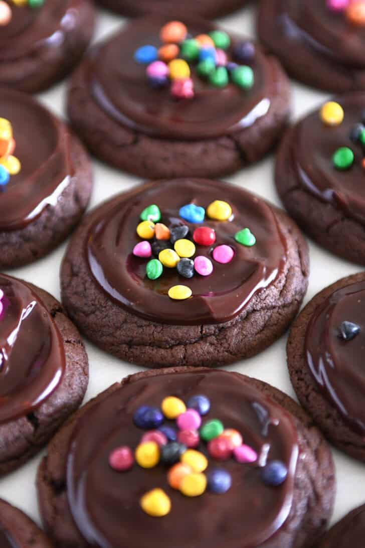 Several types of matte chocolate chip cookies on white parchment with rainbow slices on top.