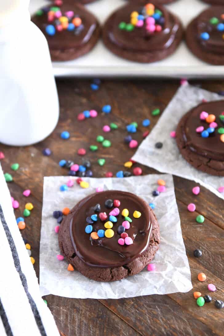Chocolate chip cookies on white parchment with ganache and rainbow chips, rainbow fries sprinkled in the background, several cookies.