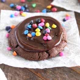 A chocolate chip cookie is spread with chocolate ganache and sprinkled with rainbow fries on white parchment.