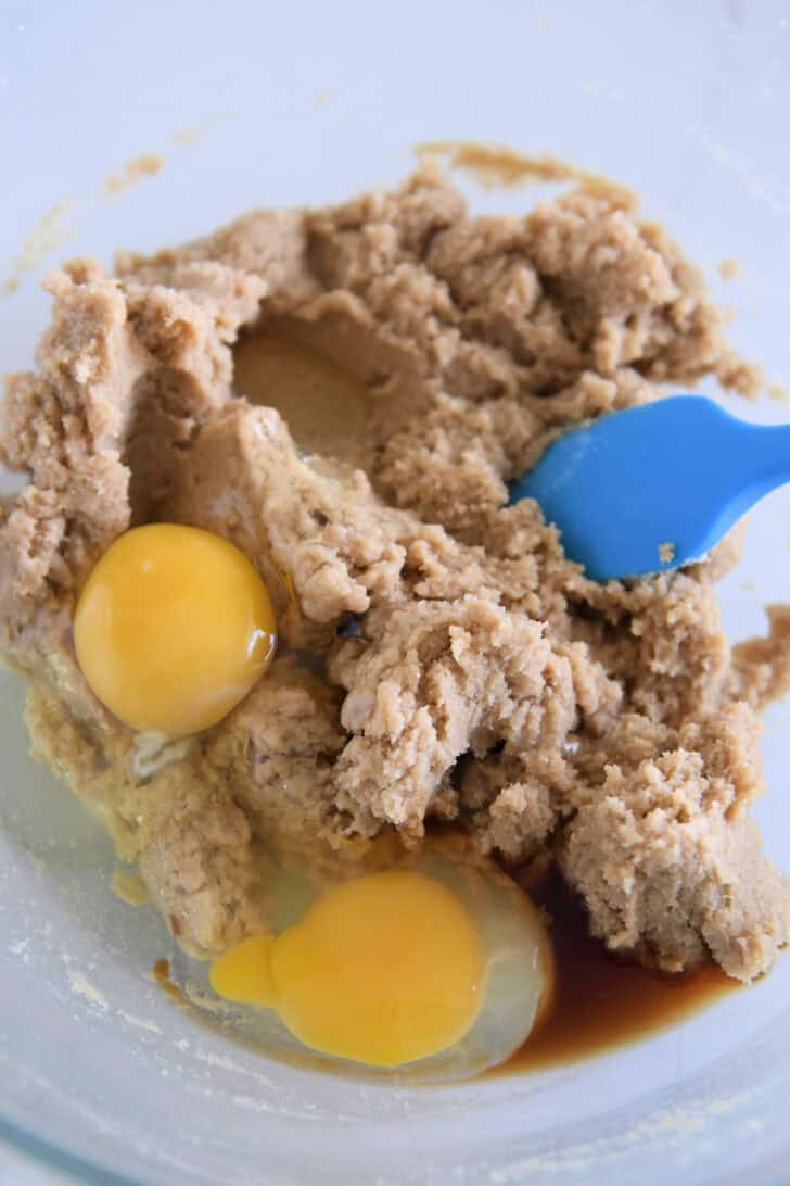 Glass bowl with two eggs and brown sugar cookie dough and blue spatula.