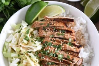 Top down photo of white bowl with chopped cabbage, white rice, grilled chicken, lime wedges, chopped cilantro and toasted sesame seeds.