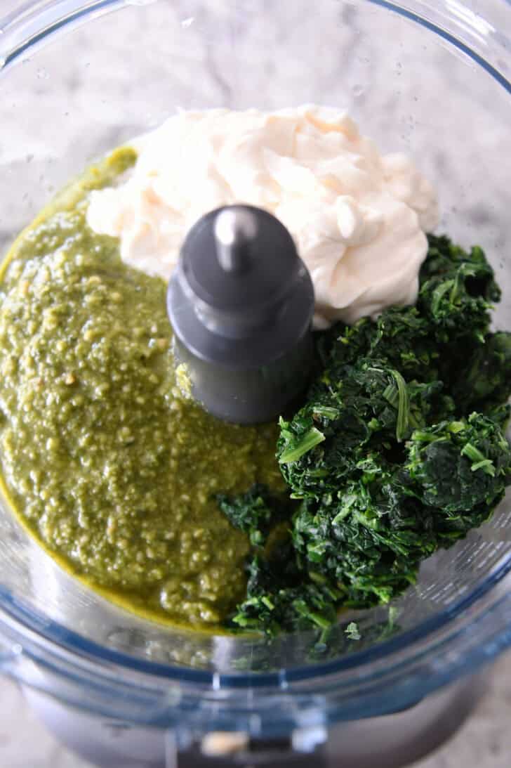Top down view of mayonnaise, pesto, and spinach in a food processor.