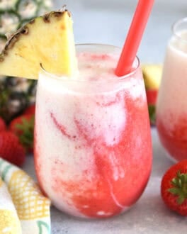 Glass cup filled with strawberry puree and icy pineapple coconut mixture with red straw and pineapple wedge on rim of glass.