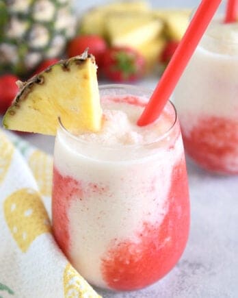 Glass cup filled with strawberry puree and icy pineapple coconut mixture with red straw and pineapple wedge on rim of glass.