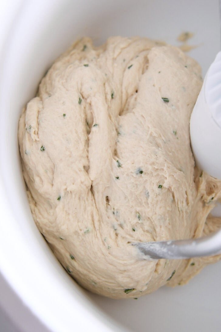 Top down view of a white mixing bowl with rosemary bread dough being mixed inside.