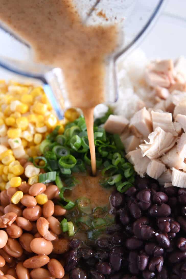 An orange sauce being poured over a bowl of chicken, beans, green onions, and corn.