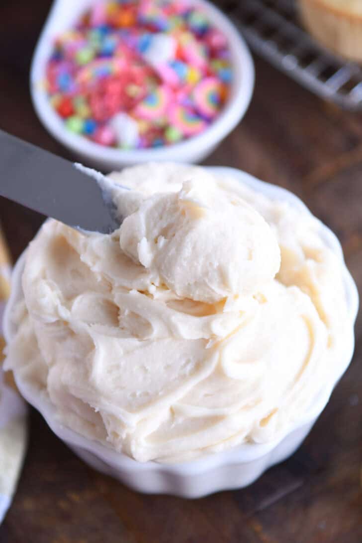 Top down view of a small white bowl with white frosting and a metal spatula taking a scoop out.