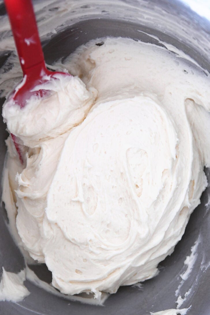 Top down view of a metal bowl with white frosting and a red spatula.