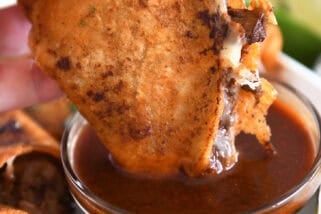 Dipping beef and cheese taco with crispy outer shell in rich birria broth in glass bowl.