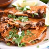 Three corn tortillas filled with beef and melty cheese topped with cilantro and onions on white platter.