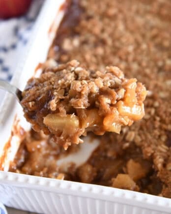 Serving spoon with heaping serving of caramel apples topped with buttery oat crisp in front of pan of caramel apple crisp.