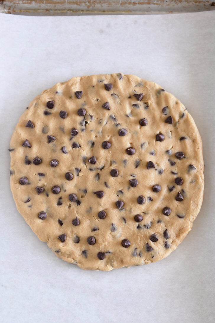 Top down view of a cookie sheet with parchment paper and a large uncooked chocolate chip cookie with extra chocolate chips sprinkled on top in the center.