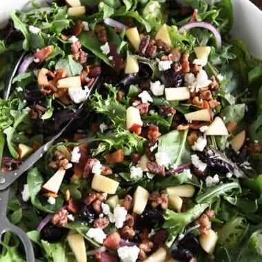 White bowl with salad tongs and mixed salad of baby greens, chopped apples, feta cheese, cooked bacon, craisins, and pumpkin seeds.