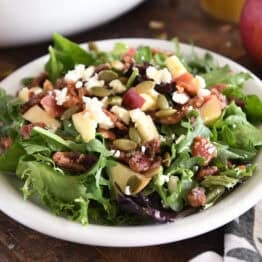 Green salad on a white plate with fried pumpkin, apples, chopped pecans, feta cheese and red onions.