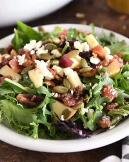 Green salad on a white plate with roasted pumpkin seeds, chopped apples, candied pecans, feta cheese and red onions.