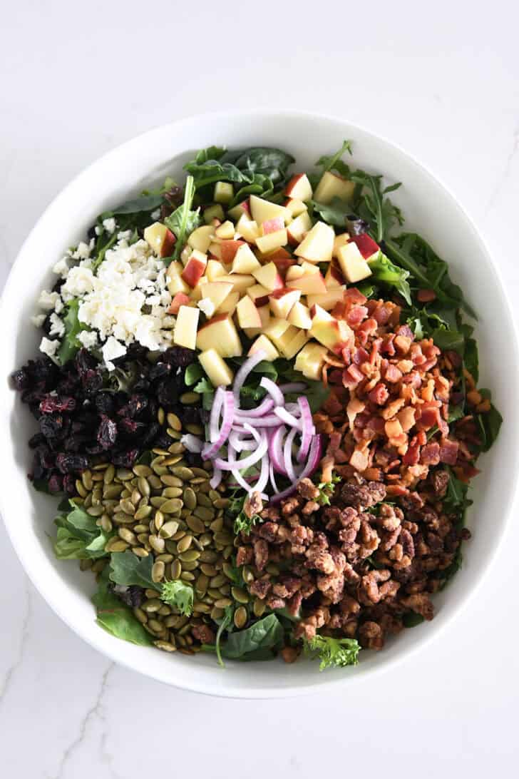 A white container with salad greens, fried seeds of thigh, targeted pecans, cooked bacon, red onion, craisins, feta.