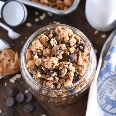 Top down view of shallow glass jar filled with peanut butter granola with roasted peanuts and dark chocolate chunks with spoonful of peanut butter to the side.