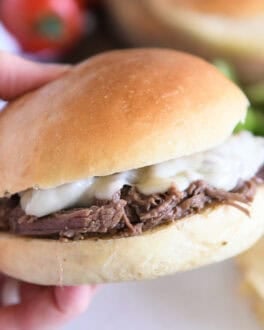 Slow cooker beef on crusty rolls with provolone cheese and one bite taken out; hand holding sandwich.