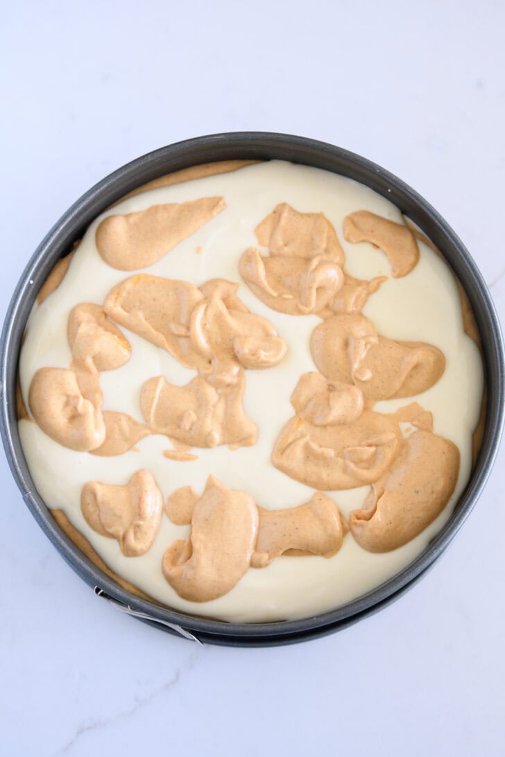 Top down view of 9-inch springform pan with dollops of pumpkin cheesecake batter on top of regular pumpkin batter.