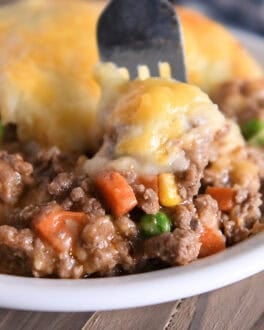 A beef and vegetable mixture topped with cheesy potatoes in a white bowl. A fork is taking a bite out.