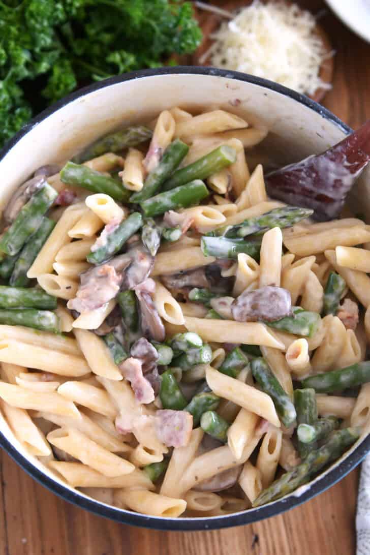 Top down view of a pot of cooked penne pasta in a creamy white sauce with chopped asparagus, and sliced mushrooms.