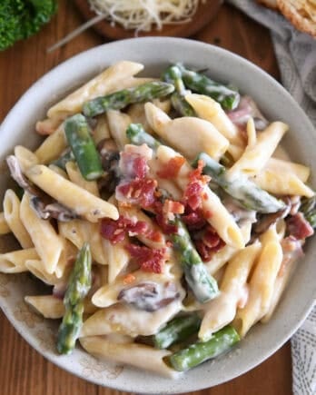 Top down view of creamy penne pasta with mushrooms, asparagus, and crumbled bacon in a white bowl.