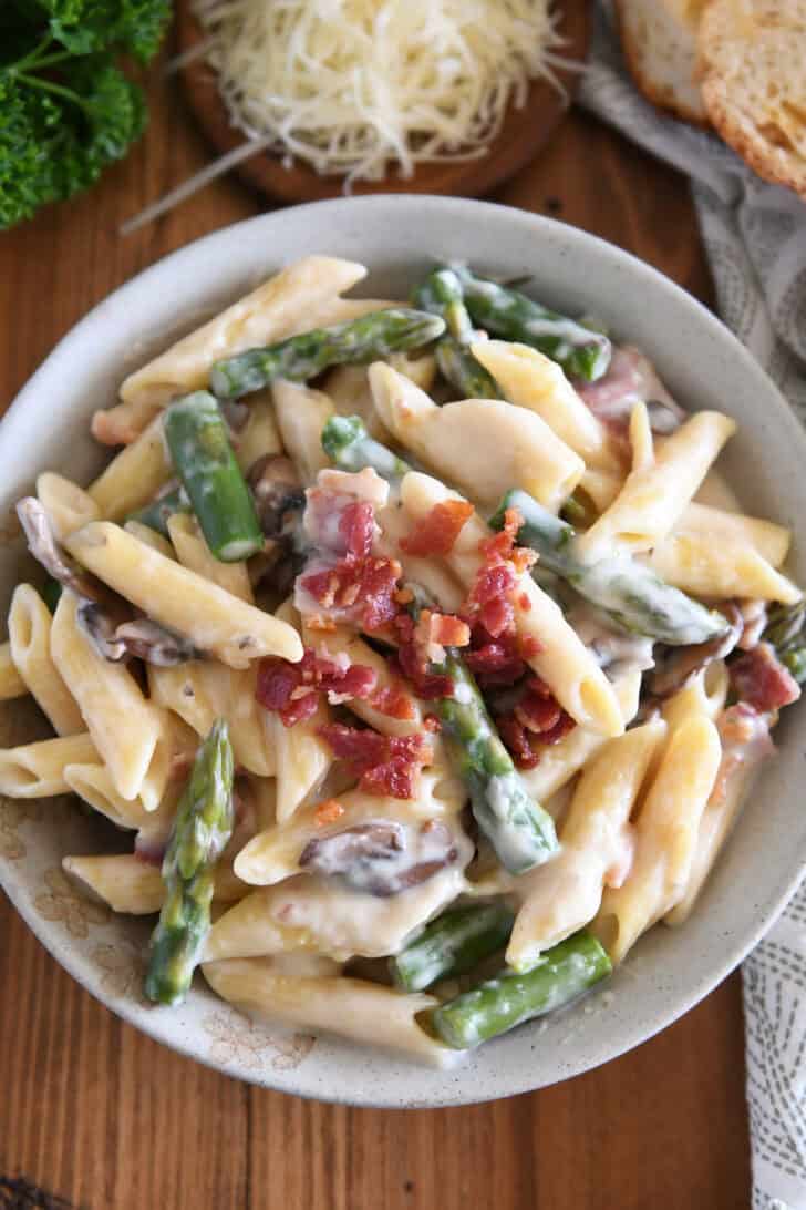 Top down view of creamy penne pasta with mushrooms, asparagus, and crumbled bacon in a white bowl.