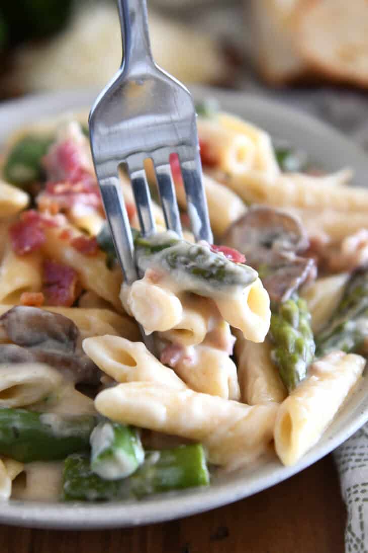 A fork taking a bite of creamy penne pasta with mushrooms, asparagus, and crumbled bacon.