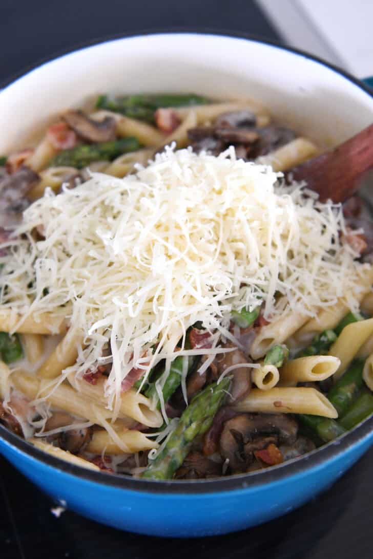 Top down view of a pot with cooked penne pasta with asparagus and sliced mushrooms topped with shredded cheese.