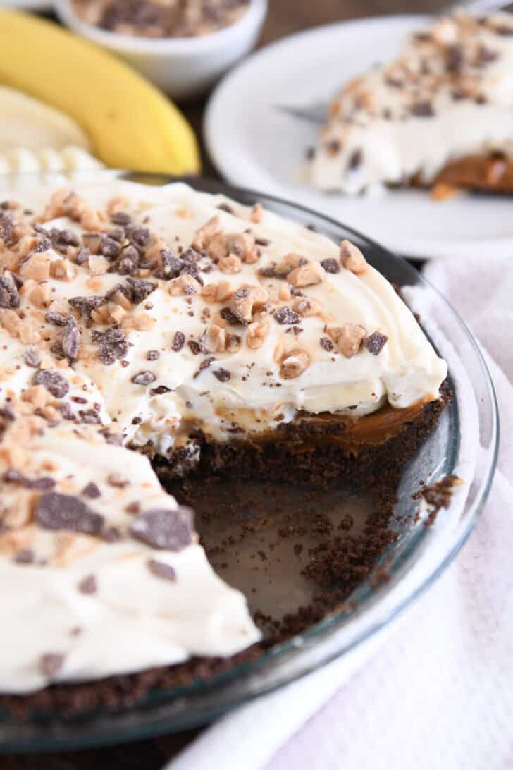 Glass 9-inch pie plate with banoffee pie with one slice removed and placed on white plate.