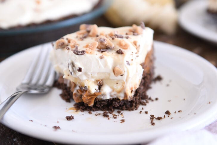 Slice of pie with chocolate cookie crust, dulce de leche, bananas, whipped cream and toffee on white plate.