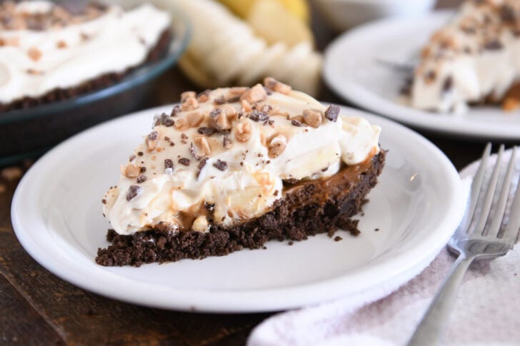 Slice of pie with chocolate cookie crust, dulce de leche, bananas, whipped cream and toffee on white plate.