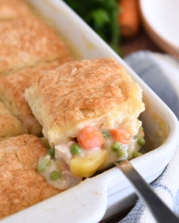 Spoon lifting out scoop of chicken pot pie filing for white 9X13-inch pan with baked biscuit on top.