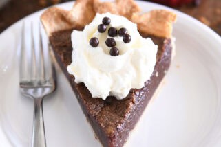 Triangle slice of chocolate chess pie on white plate with fork to the side and a dollop of whipped cream on top of pie topped with chocolate crisp pearls.