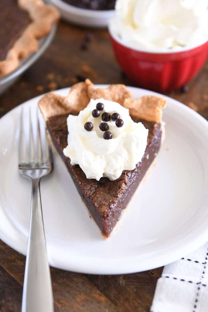 Triangle slice of chocolate chess pie on white plate with fork to the side and a dollop of whipped cream on top of pie topped with chocolate crisp pearls.