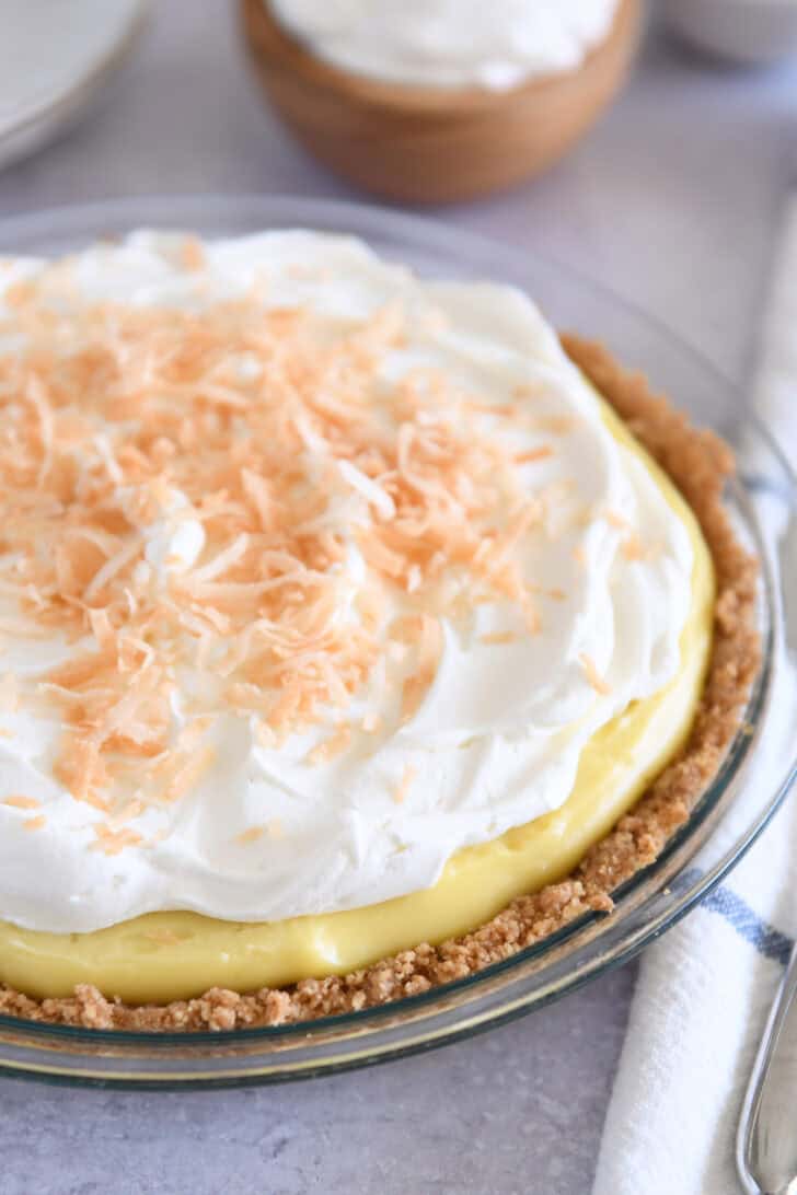 Whole coconut cream pie in glass pie plate topped with whipped cream and toasted coconut.