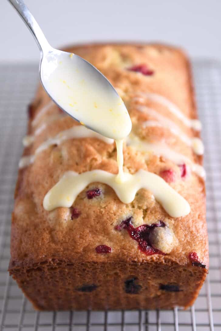 Drizzling orange glaze on cranberry orange bread with spoon.